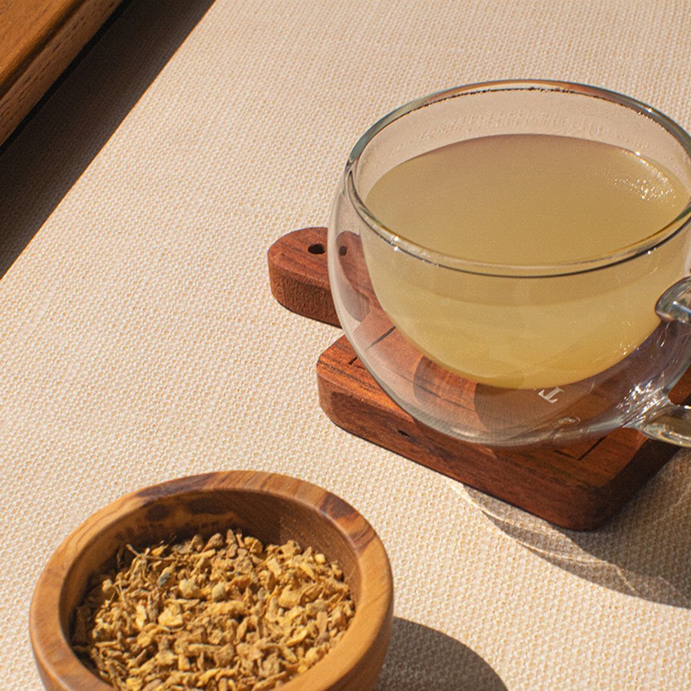 A glass cup of brewed ginger root tea placed on a wooden coaster next to a small wooden bowl of dried ginger root pieces on a beige surface.